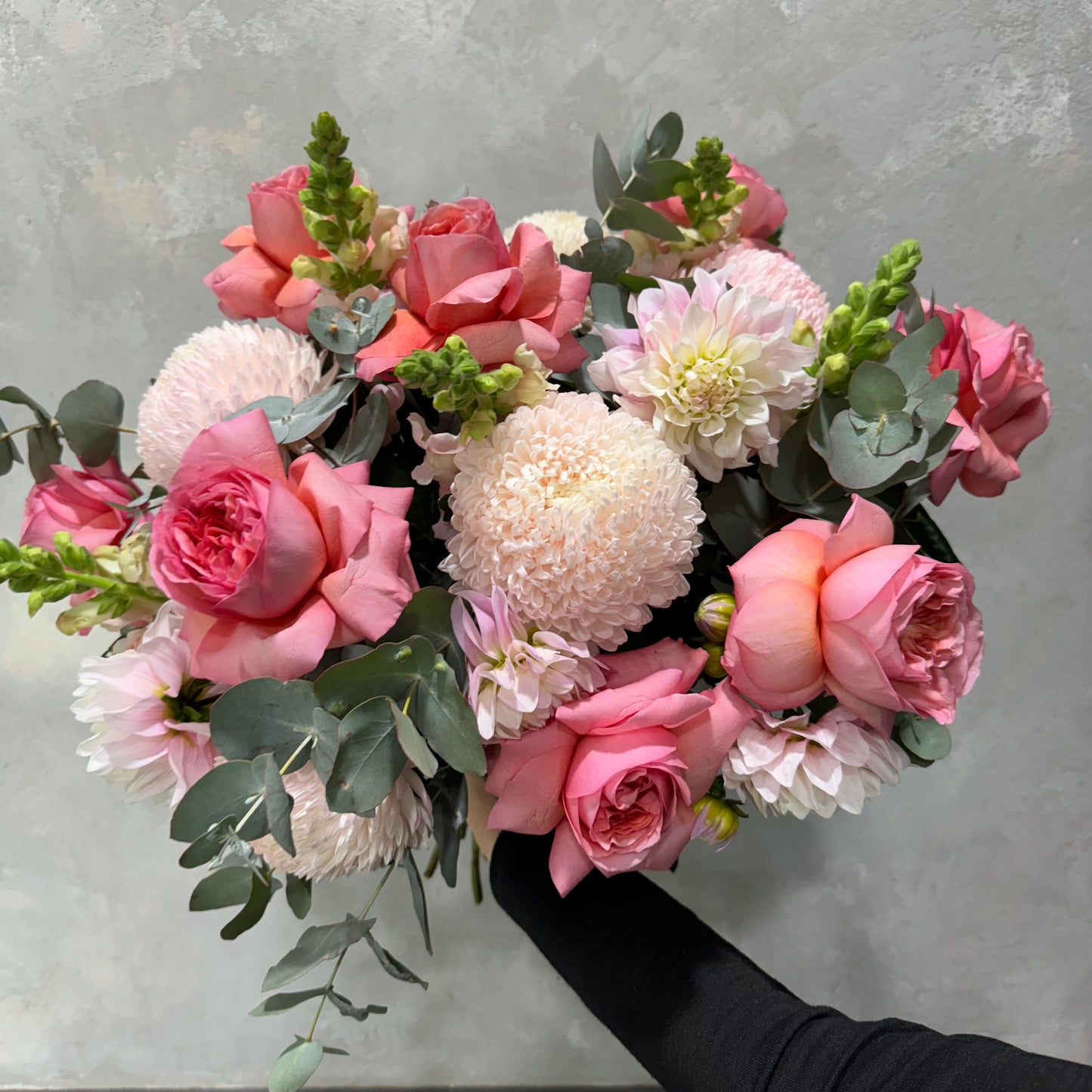 The "Pretty in Pink" bouquet by Flowers on Norton St displays seasonal flowers, highlighting pink and white roses alongside chrysanthemums against a light gray backdrop. This lush and vibrant arrangement beautifully combines various textures and soft hues.