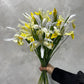 A hand holds an elegant Iris Flower Bunch by Flowers on Norton St, featuring yellow and white irises against a textured light gray background. The flowers have long, slender green stems and delicate petals. The person is wearing a black sleeve.