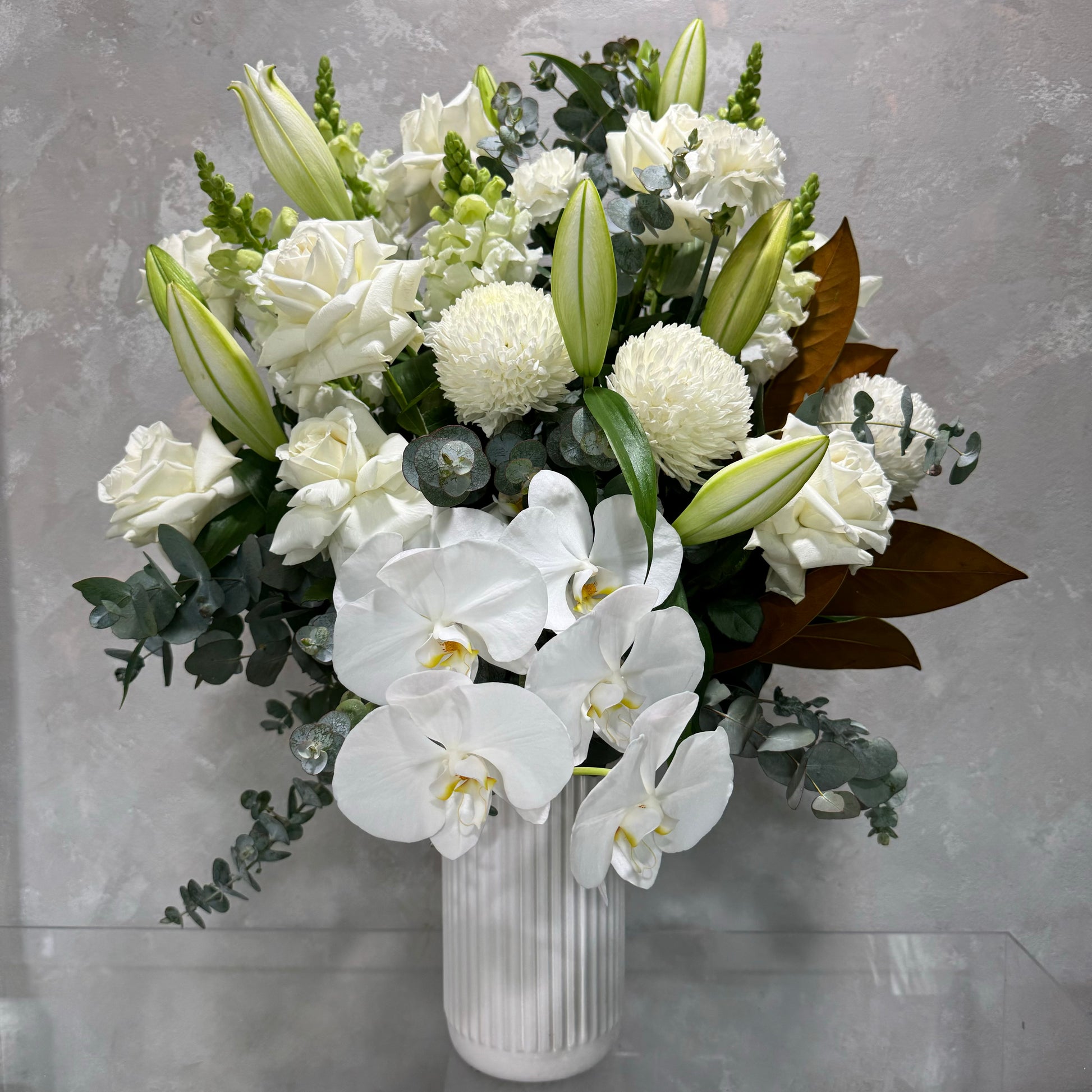 The Snowy White vase by Flowers on Norton St showcases an elegant arrangement of fresh white orchids, lilies, roses, and chrysanthemums, beautifully accented with green foliage against a textured gray background.