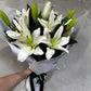 A hand holds a bouquet of FlowersonNortonSt's elegant Casablanca Lily wrapped in white paper and secured with a black ribbon. These long-lasting flowers are showcased against a light, textured wall.