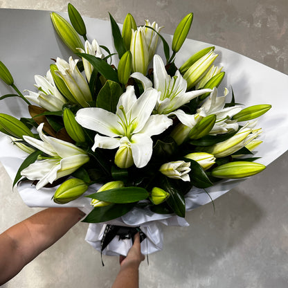 A bouquet of Casablanca Lilies, featuring closed buds and dark green leaves, wrapped in white paper, is being held against a gray background. This exquisite arrangement is crafted by FlowersonNortonSt.