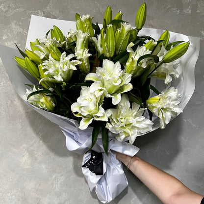 A Rose-lily Bouquet from FlowersonNortonSt, elegantly wrapped in white paper and held against a gray textured background, features fresh partially opened blooms that exude a gentle fragrance. Lush green leaves complement these exquisite flowers, enhancing their charm.