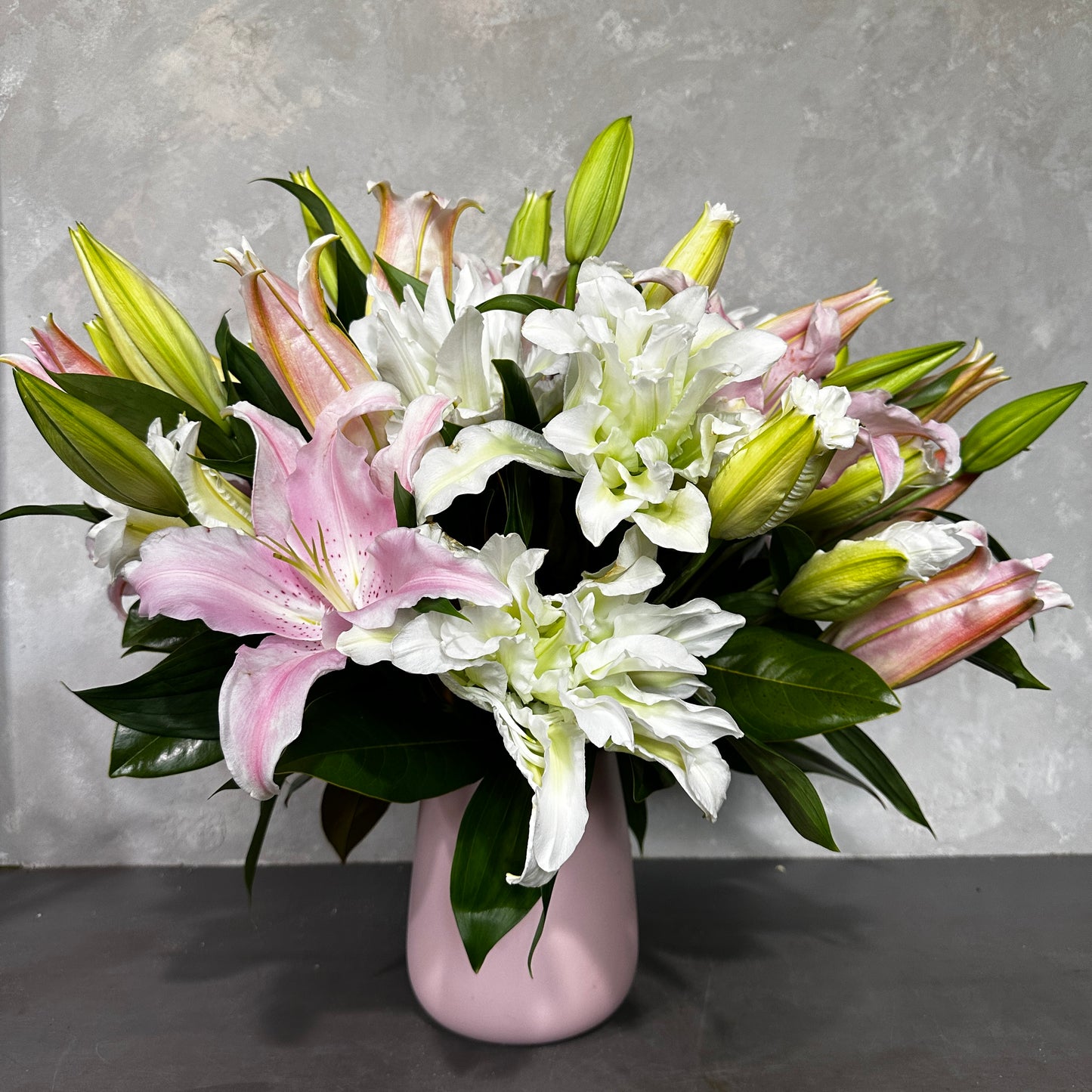 A Vase Full of Lilies from Flowers on Norton St features a bouquet of pink and white frilly rose-lilies complemented by green leaves in a pink vase, set against a textured gray background. The arrangement includes fully bloomed lilies alongside budding ones, creating a harmonious display.