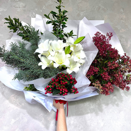 A hand holding the Style the Home - Xmas Edition bouquet from Flowers on Norton St, featuring white lilies, red berries, along with an array of vibrant green and purple foliage, elegantly wrapped in white paper tied with a red ribbon—it's the perfect festive flower arrangement against a textured gray background.