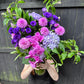 A person holds a large bouquet of jewel-toned flowers in front of their face, showcasing the "Parisian Purple" arrangement by Flowers on Norton St. It features pink dahlias, purple lisianthus, hydrangeas, and phalaenopsis orchids among lush green foliage, all set against a rustic dark wooden fence backdrop.