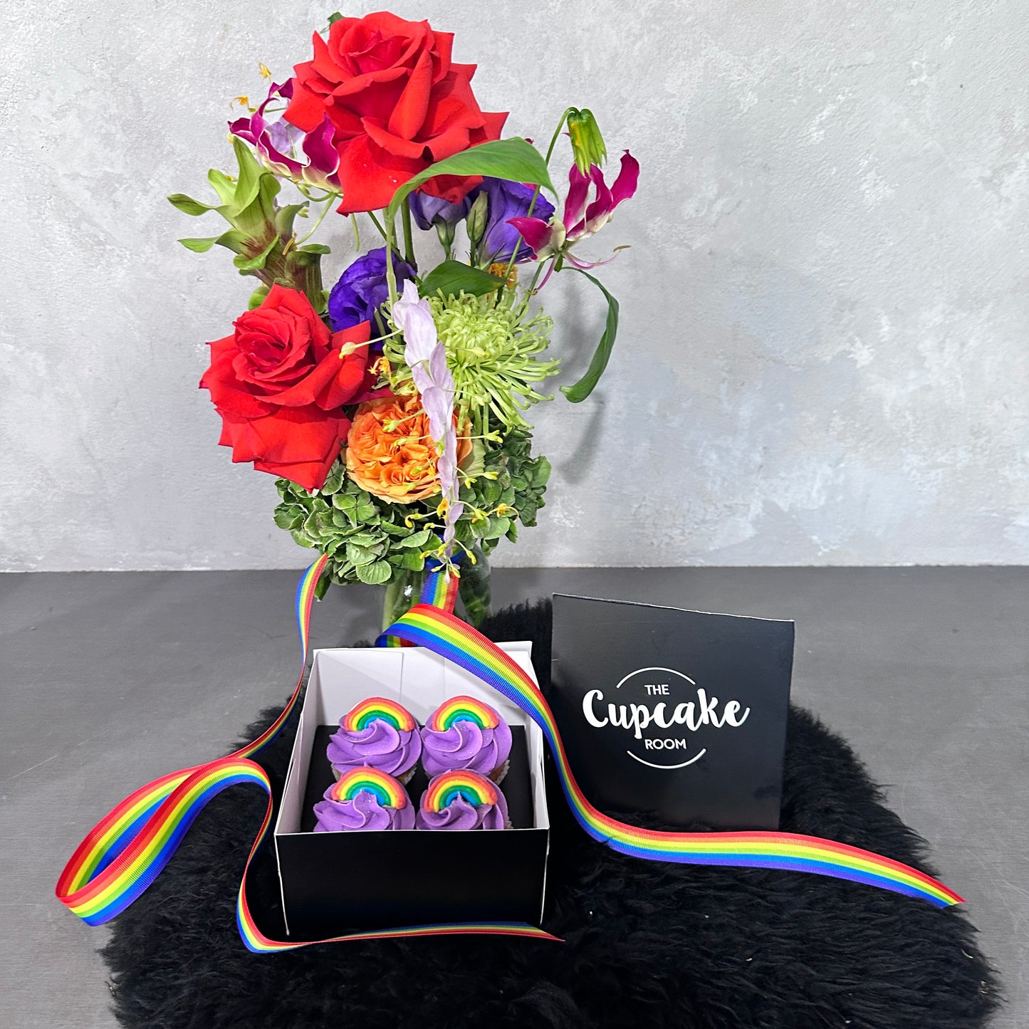 A vibrant bouquet from Flowers on Norton St featuring red, orange, and purple flowers in a vase is paired with Rainbow Pride cupcakes, topped with colorful frosting to celebrate Pride Month. A rainbow ribbon elegantly wraps around the cupcake box on a fluffy black surface, accompanied by a card that reads "The Cupcake Room.