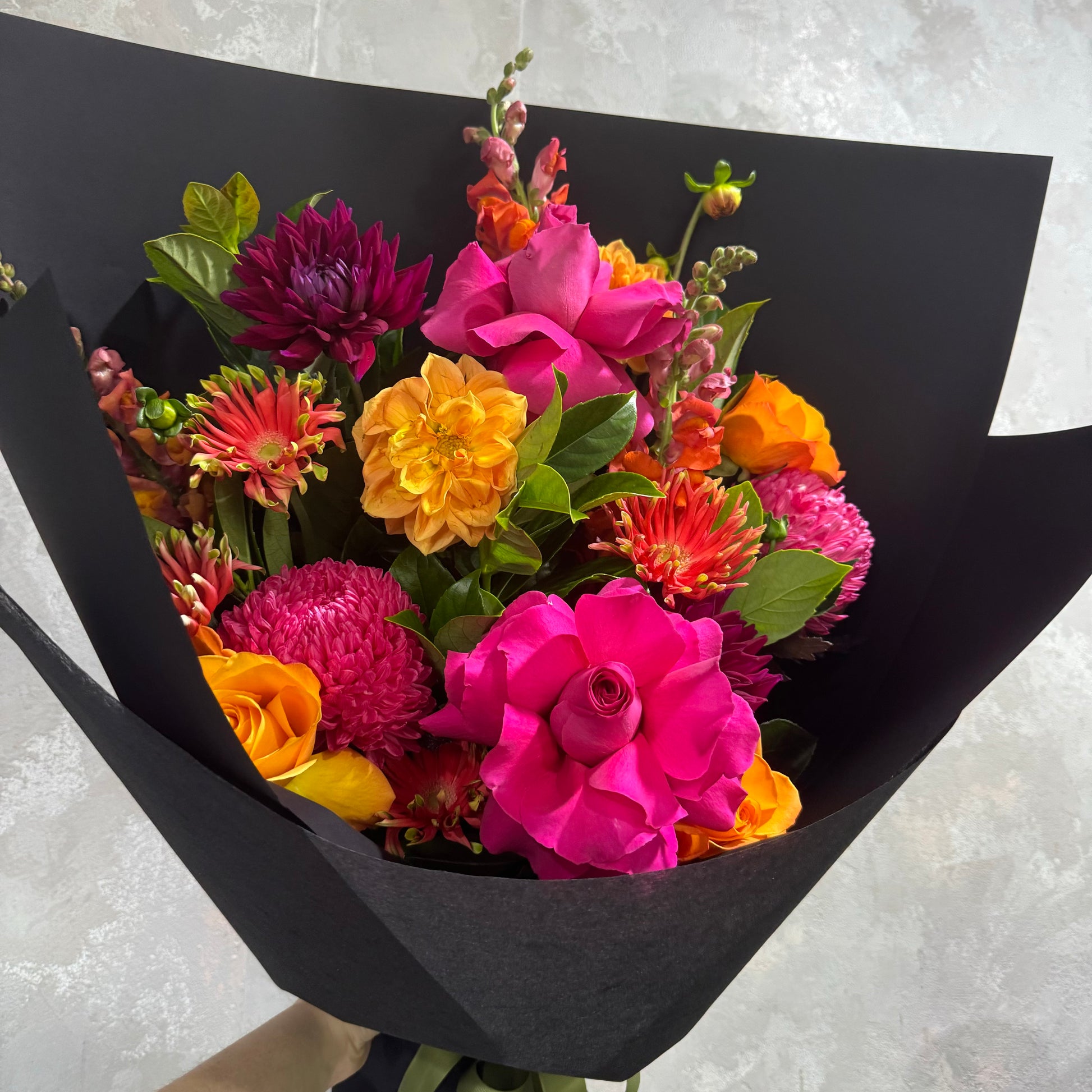 The "Summer Pop" bouquet by Flowers on Norton St features a vibrant mix of colorful blooms, including pink roses, orange and yellow dahlias, magenta chrysanthemums, and green foliage, all elegantly wrapped in black paper. This captivating arrangement stands out beautifully against a light gray textured background.