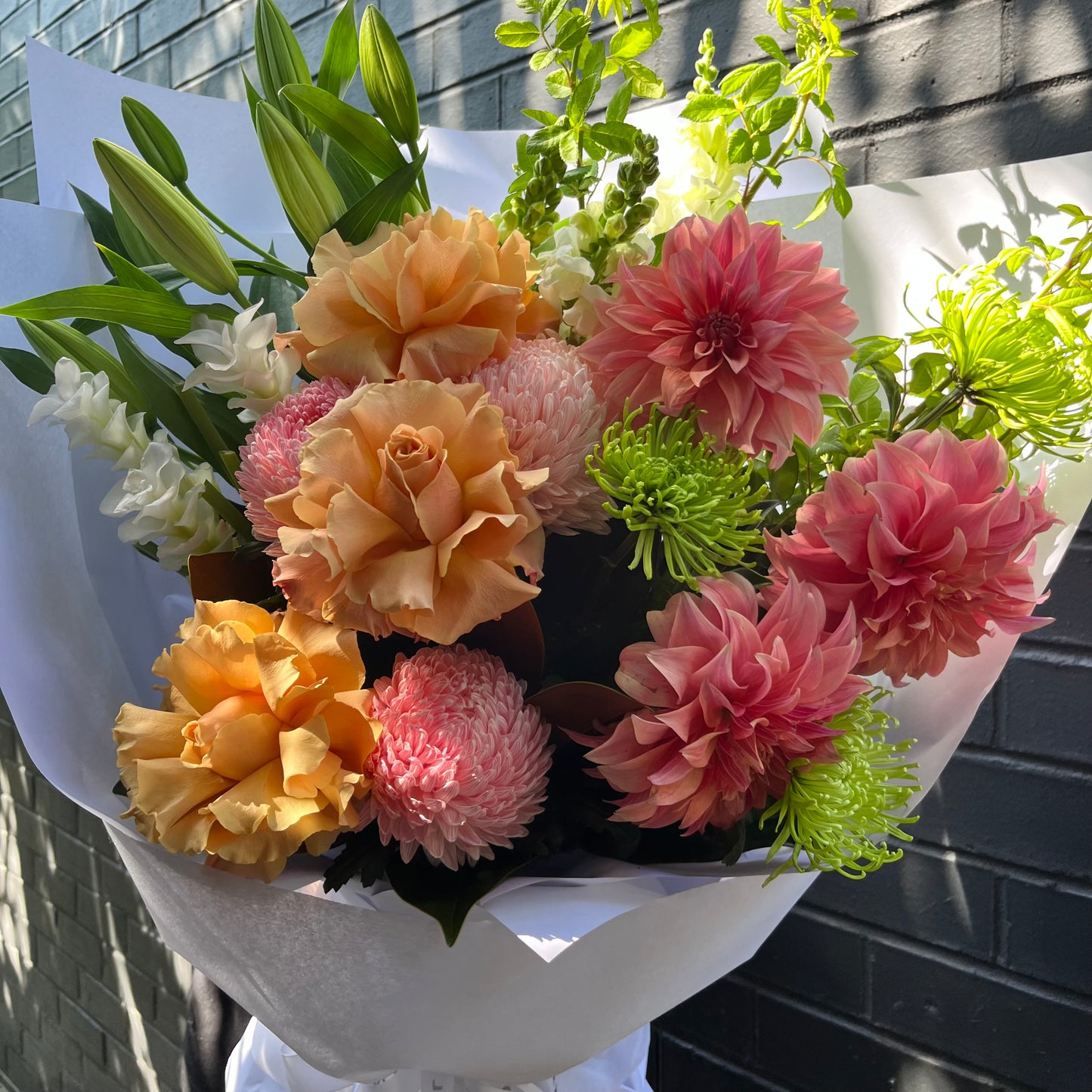 A vibrant bouquet named "Floral Coral" by Flowers on Norton St showcases lush roses, pink dahlias, white lilies, and green chrysanthemums elegantly wrapped in white paper. The arrangement is beautifully displayed against a sunlit brick wall backdrop.