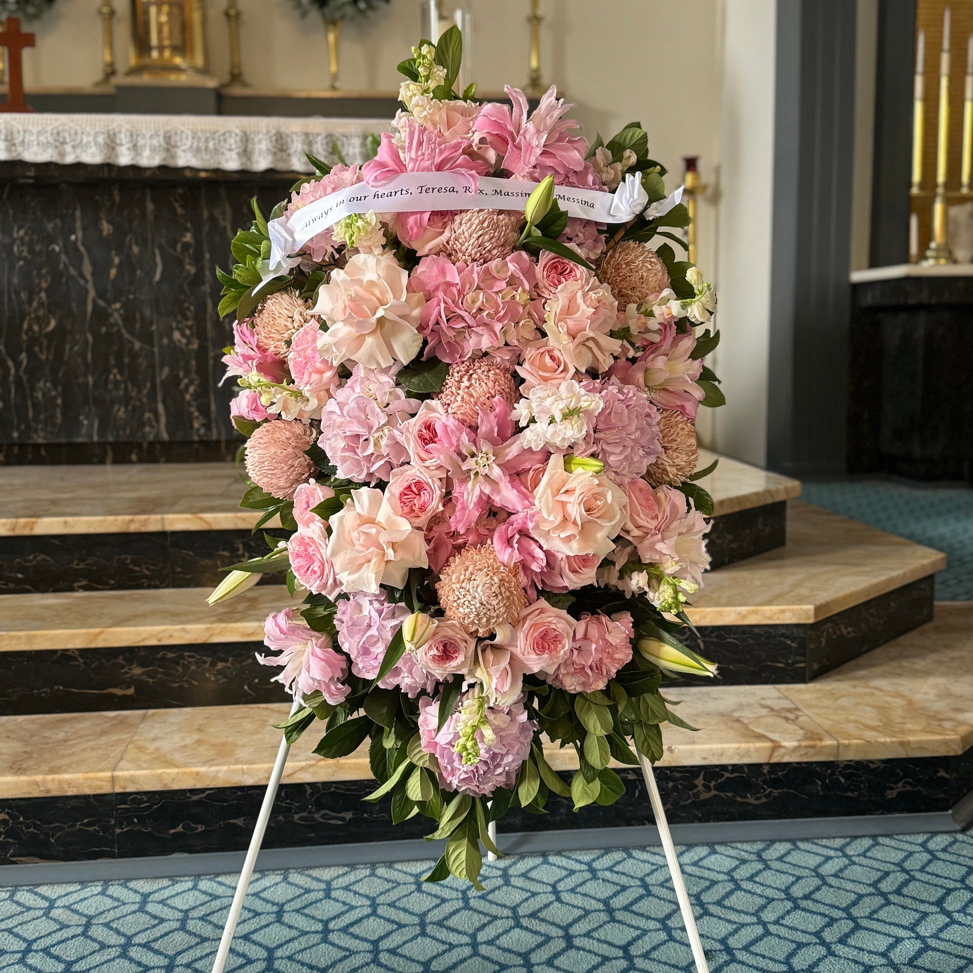 The Sympathy Hanging Sheaf by FlowersonNortonSt, featuring pink and peach flowers like roses and mums, drapes across a white easel in a church. This heartfelt tribute stands before a marble altar on blue carpet and is adorned with a condolence ribbon.