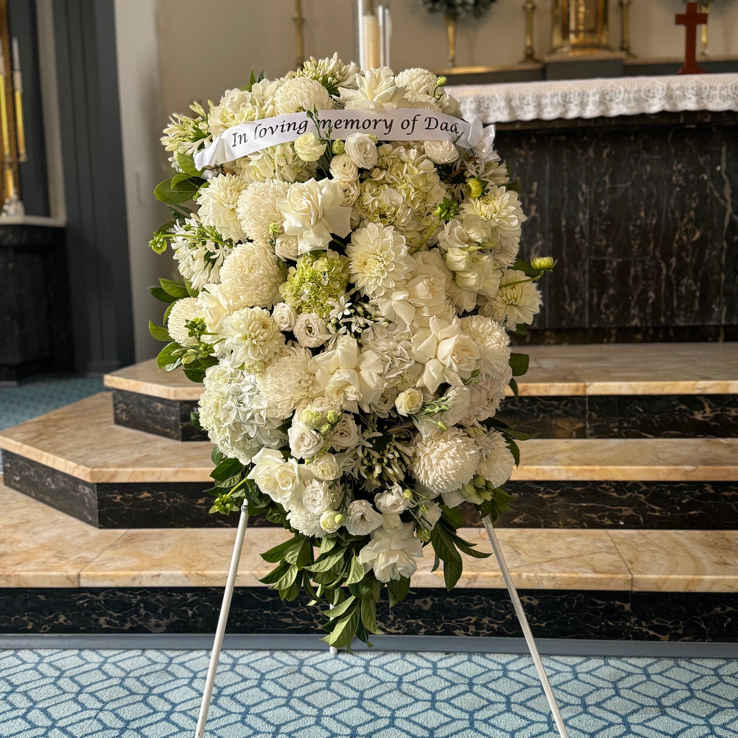 The "Sympathy Hanging Sheaf" by FlowersonNortonSt, featuring white roses and chrysanthemums, is displayed on a tripod in a church. A ribbon reading "In loving memory of Dad" adorns the top, with marble altar steps visible in the background for those gathered in remembrance.