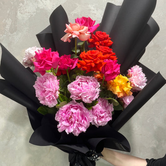 An eye-catching Peony + Rose Bouquet from Flowers on Norton St, featuring a festive mix of vibrant pink, red, orange, and yellow roses wrapped in elegant black paper. The stunning contrast between the colorful flowers and the dark wrap evokes an image reminiscent of a bouquet against a gray backdrop.