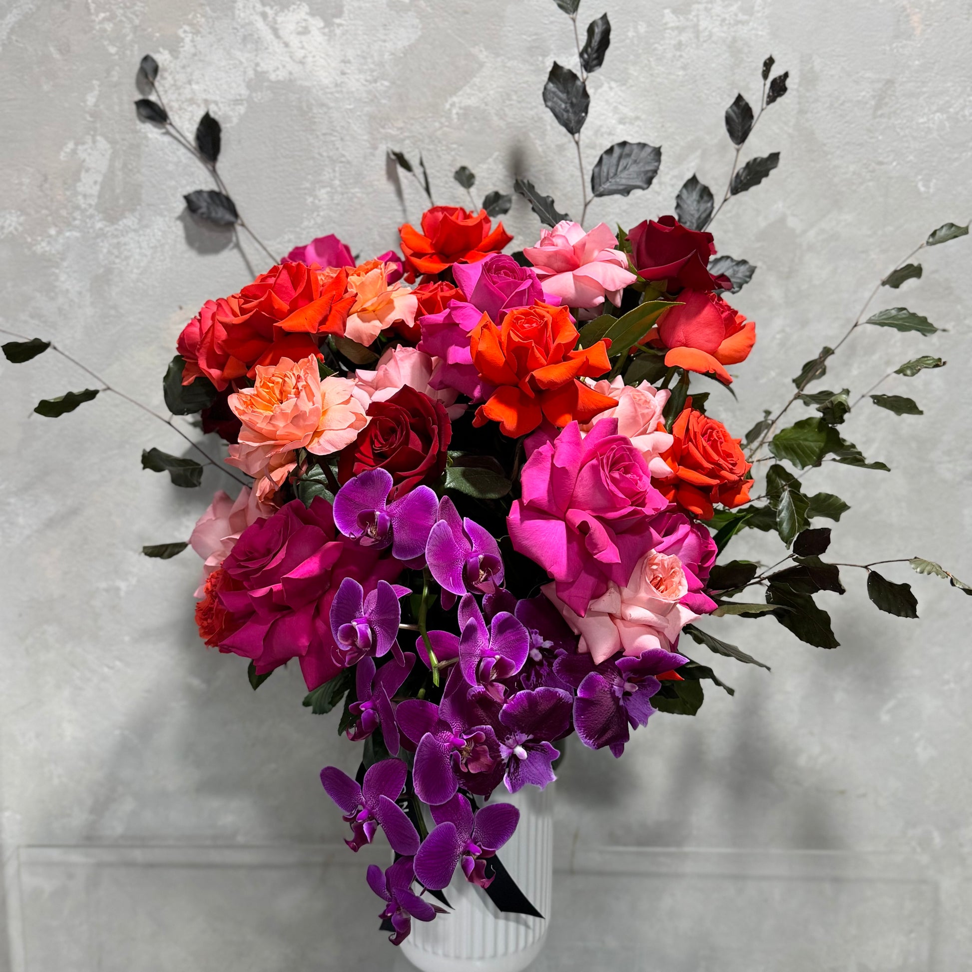 The Colombian Rose Extravaganza Arrangement by Flowers on Norton St is a vibrant bouquet in a white vase with pink, orange, and Colombian red roses and cascading Phalaenopsis orchids. Dark green leaves add contrast against a textured gray background.