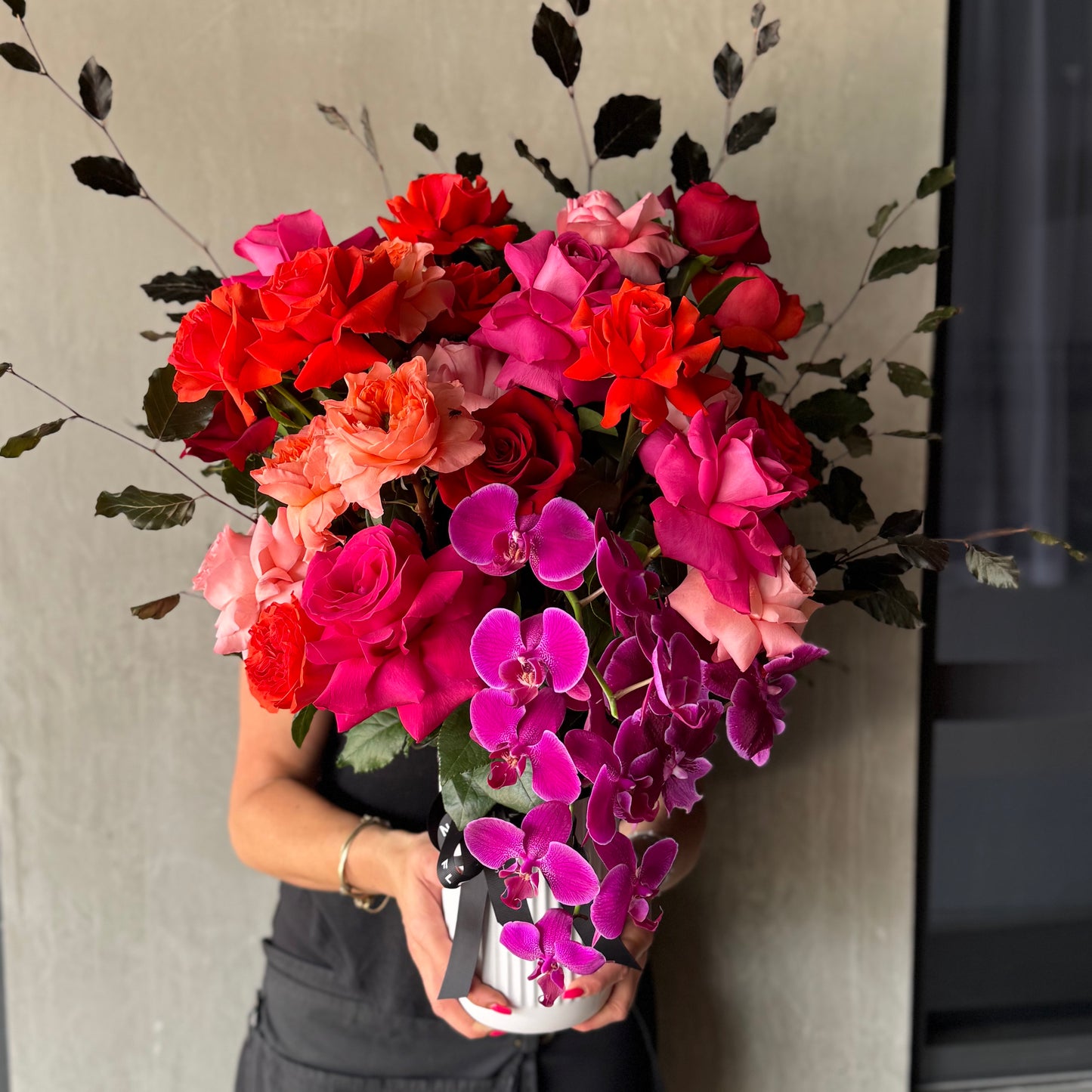 A person holding the Colombian Rose Extravaganza Arrangement from Flowers on Norton St, featuring vibrant roses and elegant pink Phalaenopsis orchids, set against a neutral background.