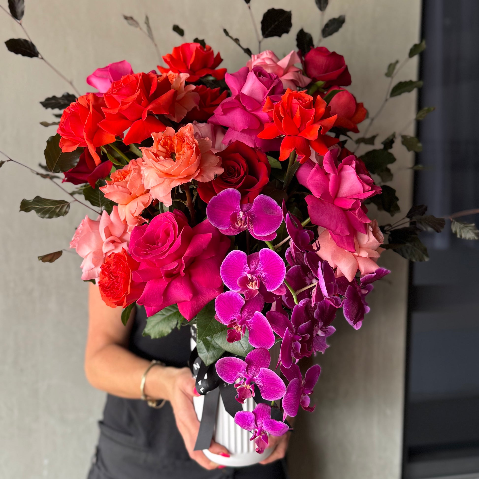 A person partially obscured in black holds the Colombian Rose Extravaganza Arrangement by Flowers on Norton St, featuring vibrant Colombian Roses and bright purple Phalaenopsis orchids in a white vase, surrounded by lush dark green leaves to create a stunning floral centerpiece.