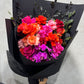 The Colombian Rose Extravaganza Arrangement by Flowers on Norton St is a vibrant mix of red, orange, pink, and fuchsia Colombian roses with bright purple Phalaenopsis orchids and dark foliage, elegantly wrapped in black paper.