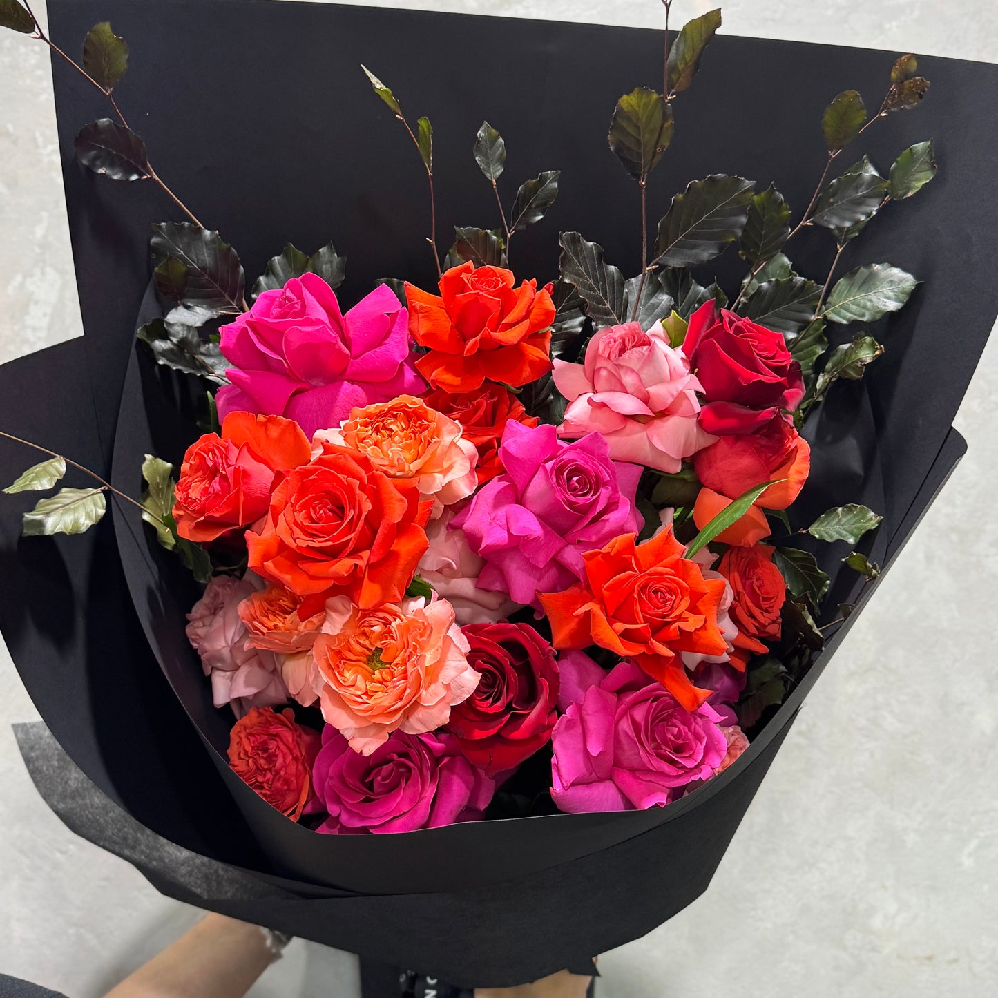 The Colombian Rose Extravaganza Arrangement by Flowers on Norton St features vibrant Colombian roses in pink, orange, and red shades, beautifully wrapped in black paper with decorative dark leaves.