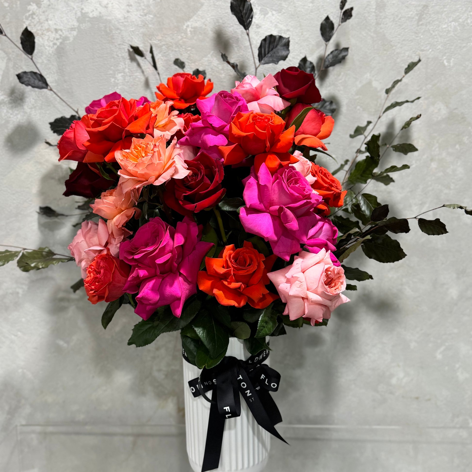 The Colombian Rose Extravaganza Arrangement by Flowers on Norton St features a vivid mix of pink, red, and orange roses in a white vase with a black ribbon against a textured gray background.