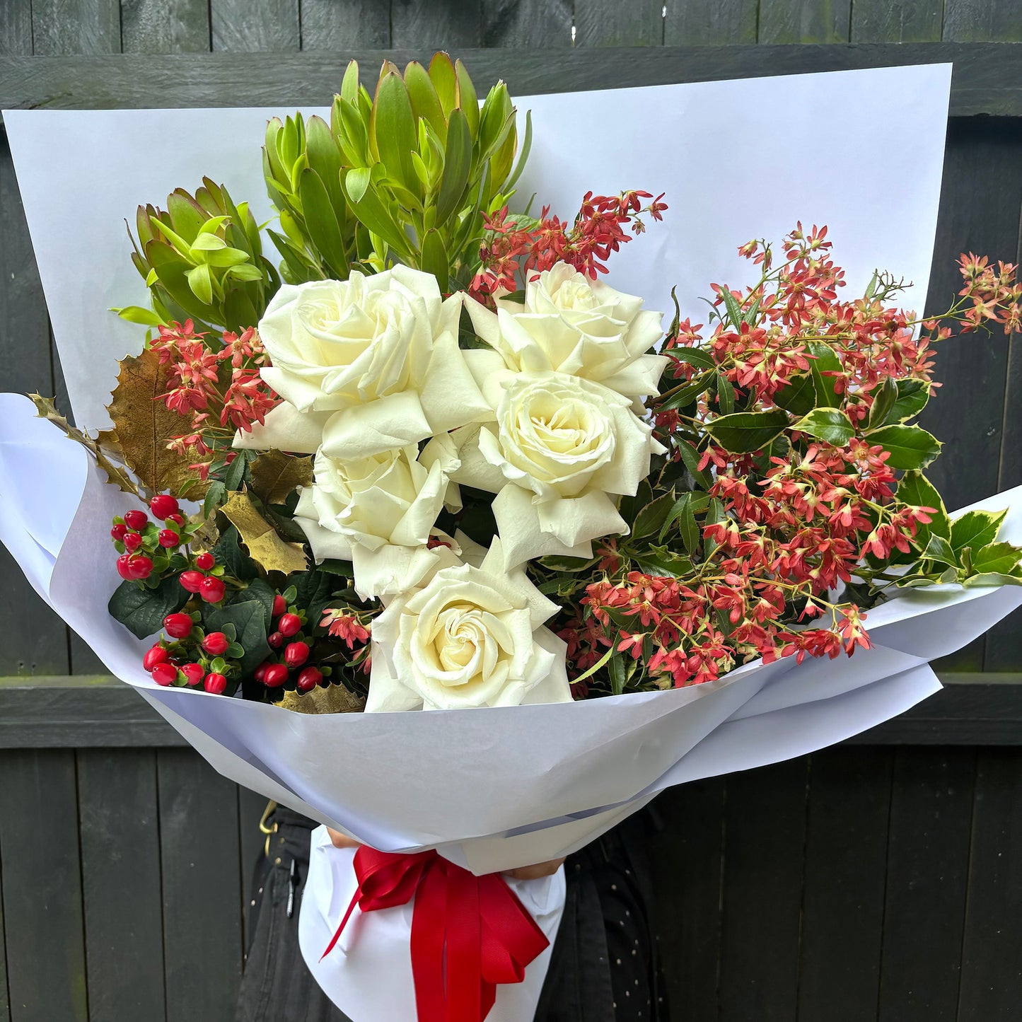 A bouquet consisting of white roses, red berries, and green foliage, accented with sprigs of holly, is wrapped in elegant white paper and tied with a striking red ribbon. It rests beautifully against a dark wooden surface. This creation is known as "Jingle all the way" by Flowers on Norton St.