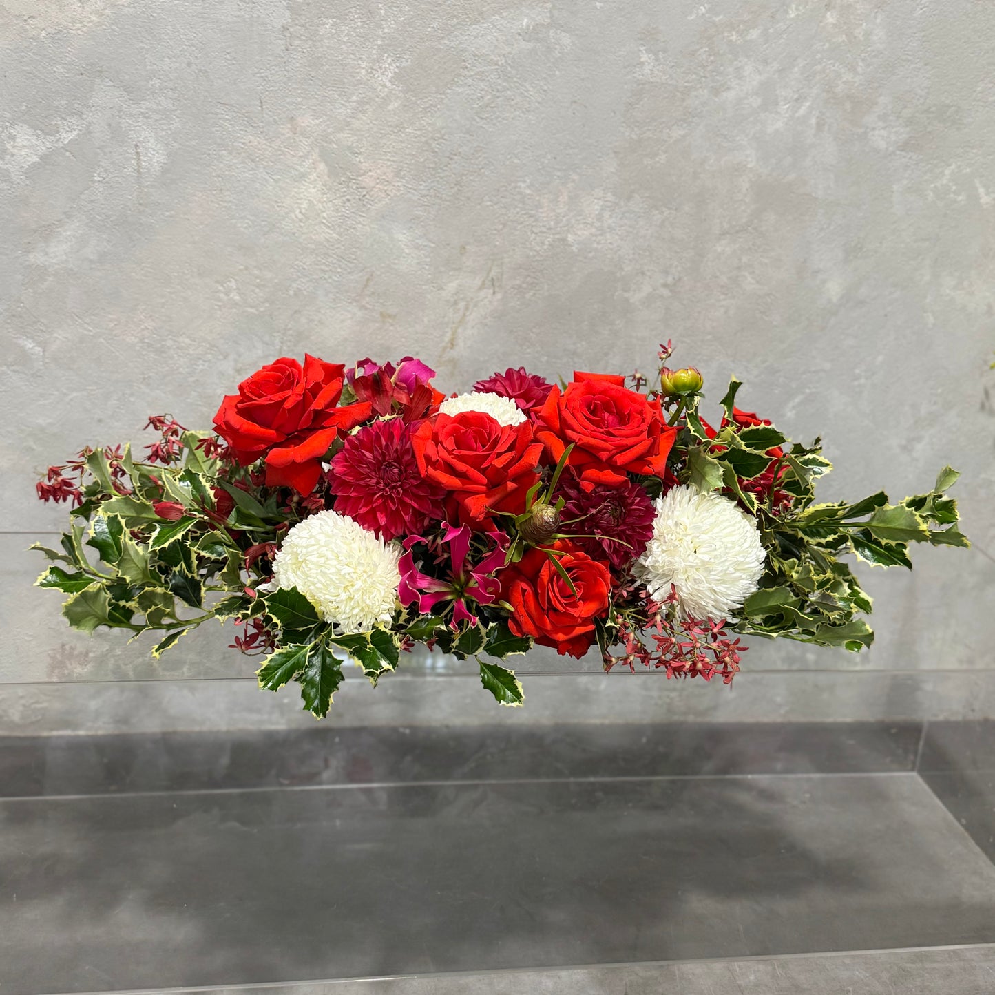 Introducing the Jolly Long + Low Centrepiece by Flowers on Norton St, showcasing an elegant floral arrangement of red roses, fuchsia flowers, and white blooms surrounded by festive greenery. This exquisite piece is set against a light gray textured background, making it the perfect addition to your Christmas table decor.