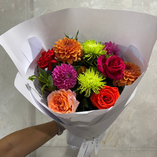 The Little Pop of Colour Posy Bouquet by FlowersonNortonSt features florists' favorites like dahlias and seasonal blooms in red, orange, pink, and green. It's wrapped in white paper and held by a person with a watch against a plain gray wall.