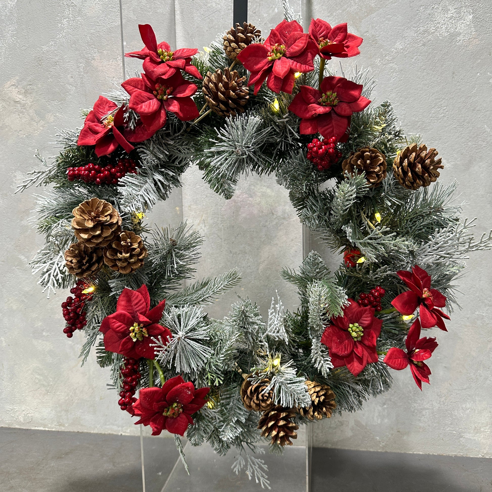 A Mistletoe Wreath by Flowers on Norton St, featuring artificial blooms, pine cones, red poinsettia flowers, and green pine branches against a gray background, beautifully captures the festive season's spirit.