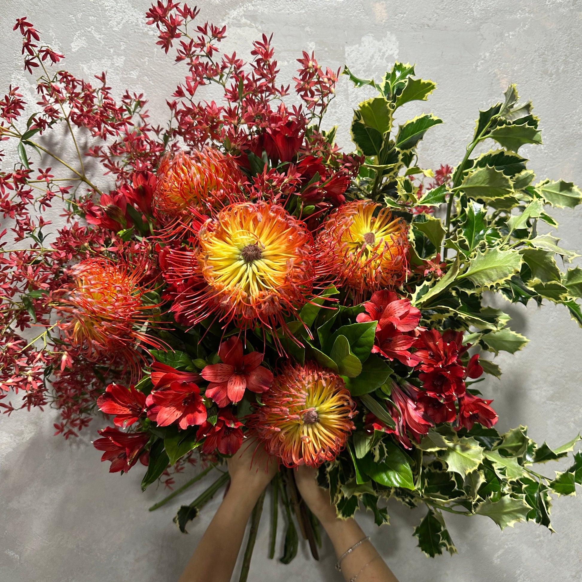 A pair of hands holds up the Native Christmas Bouquet from Flowers on Norton St, showcasing an elegant arrangement of large yellow and red protea flowers, complemented by rich red blooms and lush green foliage, against a textured gray background. This bouquet adds the perfect touch for an elegant holiday decoration.