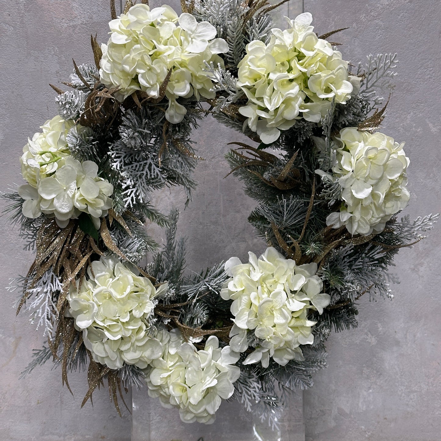 Introducing the Noel Wreath by Flowers on Norton St, a festive decoration adorned with white hydrangea flowers and frosted greenery, beautifully accented with dried gold Christmas foliage, set against a gray backdrop.