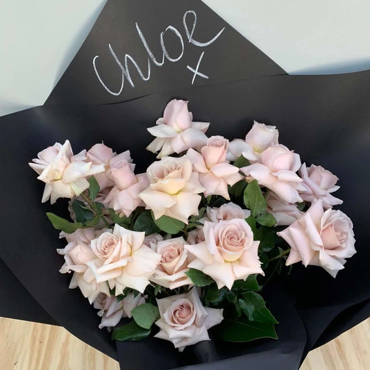 The Personalised Rose Bouquet by Flowers on Norton St showcases pale pink roses meticulously wrapped in black paper, with "Chloe x" artfully inscribed on one side. This elegant gift sits gracefully on a light wooden surface, perfectly combining sophistication and charm.