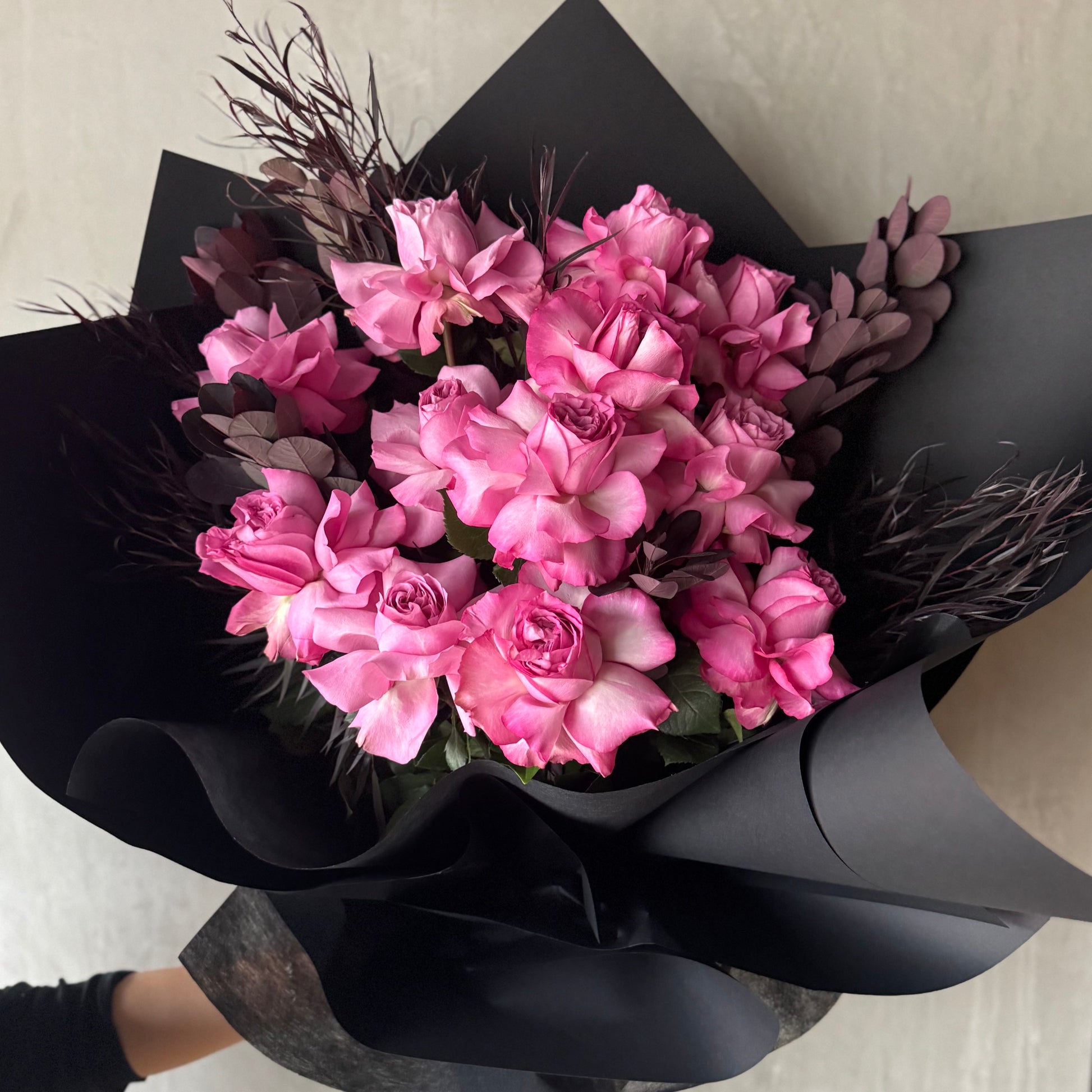 Flowers on Norton St's Queens Crown Rose Bouquet showcases vibrant pink and mauve roses wrapped in black paper. The chocolate foliage adds depth, highlighting the sophisticated look of this exquisite arrangement against a light background.