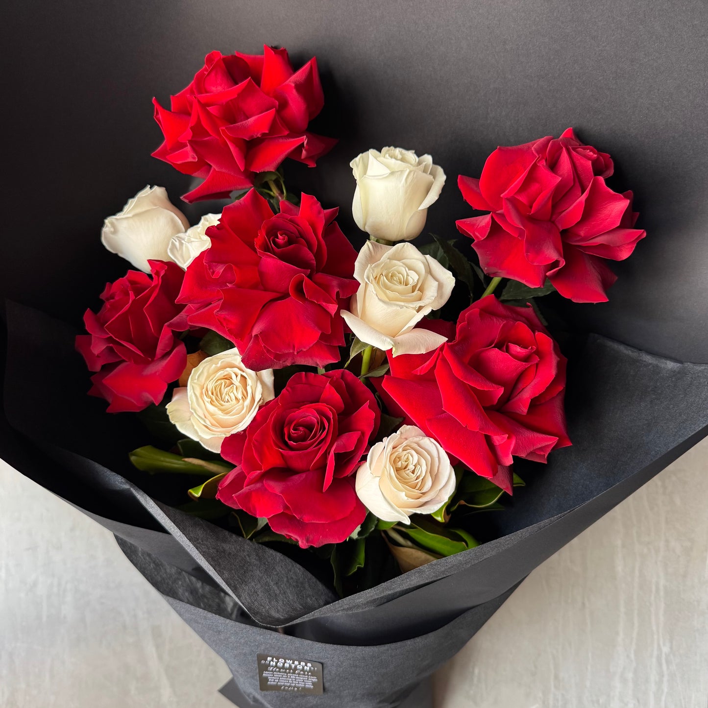 A bouquet wrapped in black paper featuring vibrant red and white roses. The Flowers on Norton St's 12-rose Red and White Bud + Bloom Rose Bouquet elegantly contrasts rich red blooms with soft white ones for a striking display.