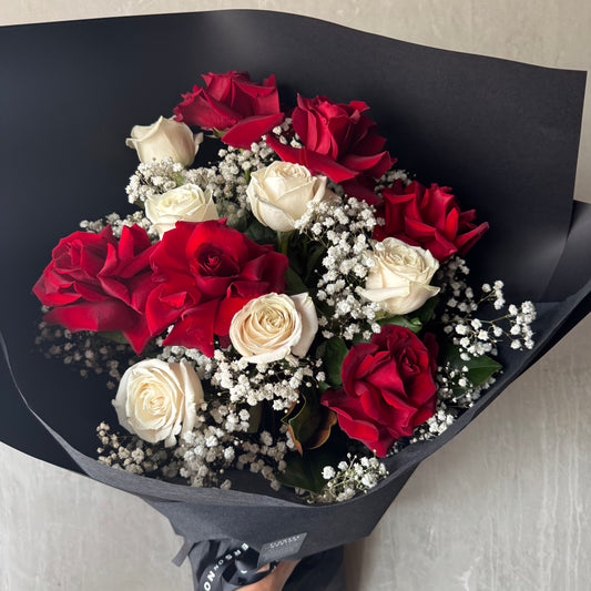 Flowers on Norton St's Bud + Bloom Rose Bouquet with Babies Breath features a 12-rose bouquet of red and white roses elegantly accented by baby's breath, wrapped in sleek black paper against a gray backdrop.