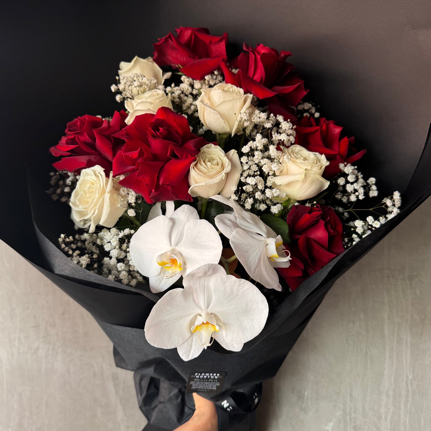 The Red and White Bud + Bloom Rose Bouquet by Flowers on Norton St features a stunning mix of red and white roses, delicate orchids, and baby's breath, elegantly wrapped in black paper.