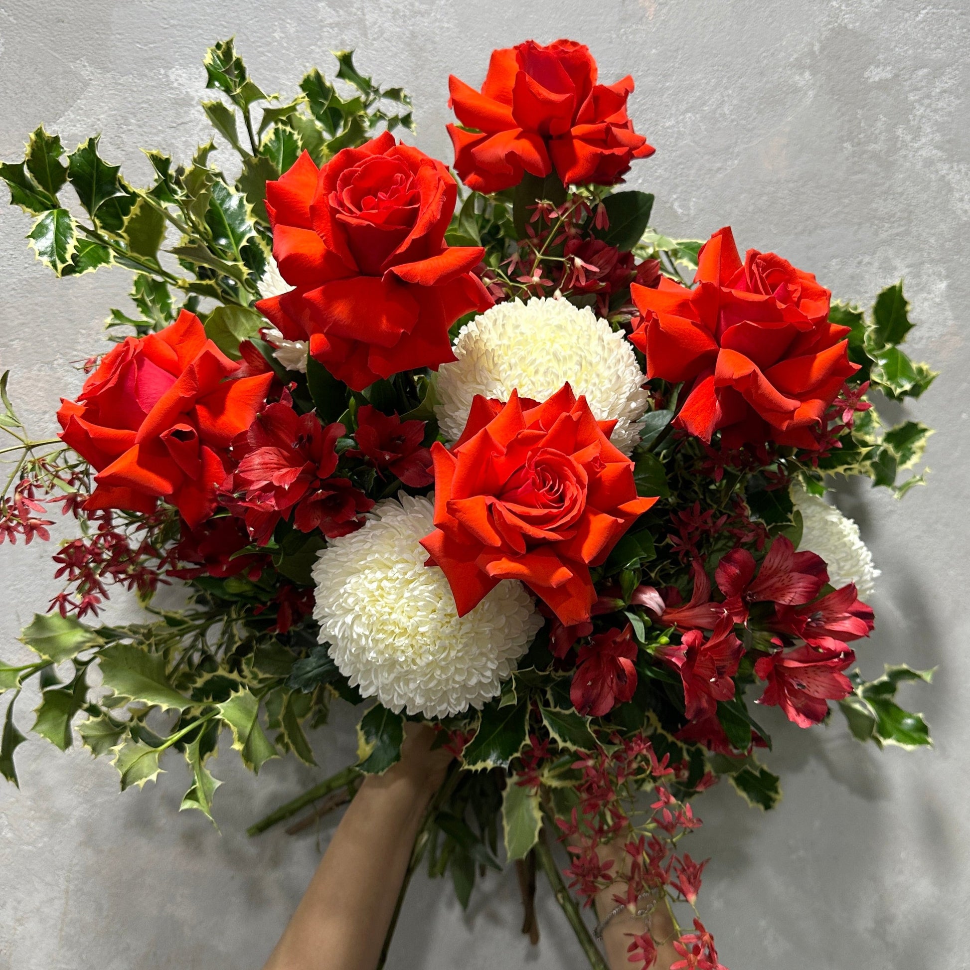 Introducing the "Rose & Holly Delight" by Flowers on Norton St, a vibrant bouquet of red roses, white chrysanthemums, and assorted greenery. This festive floral table centerpiece is expertly crafted and elegantly displayed against a neutral background.