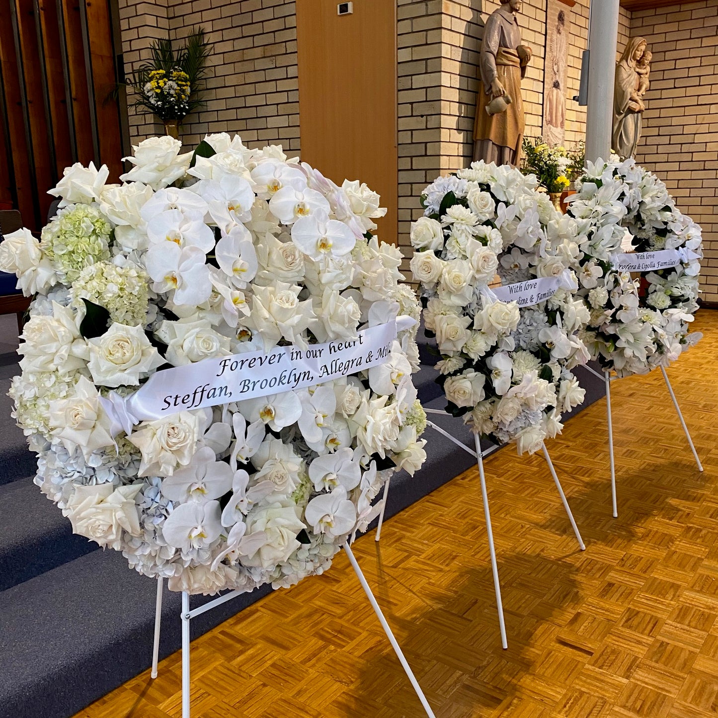 Three large floral wreaths from FlowersonNortonSt, featuring white roses and orchids on stands in a church setting. The first wreath is adorned with a ribbon bearing the message, "Forever in our hearts Steffan, Brooklyn, Allegra & Nia," with statues visible in the background.