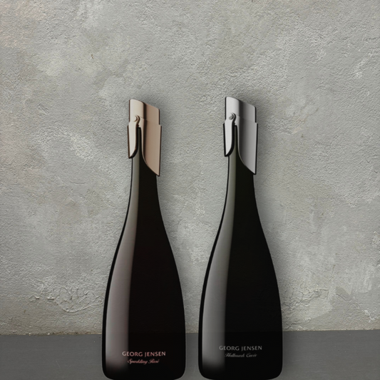 Two elegant, dark wine bottles against a textured gray background. Each bottle features a distinctive metallic accent on the neck—one gold, the other silver. Subtle text highlights their labels: "Flowers on Norton St Georg Jensen Sparkling Wine" and "Georg Jensen Mulled Wine.