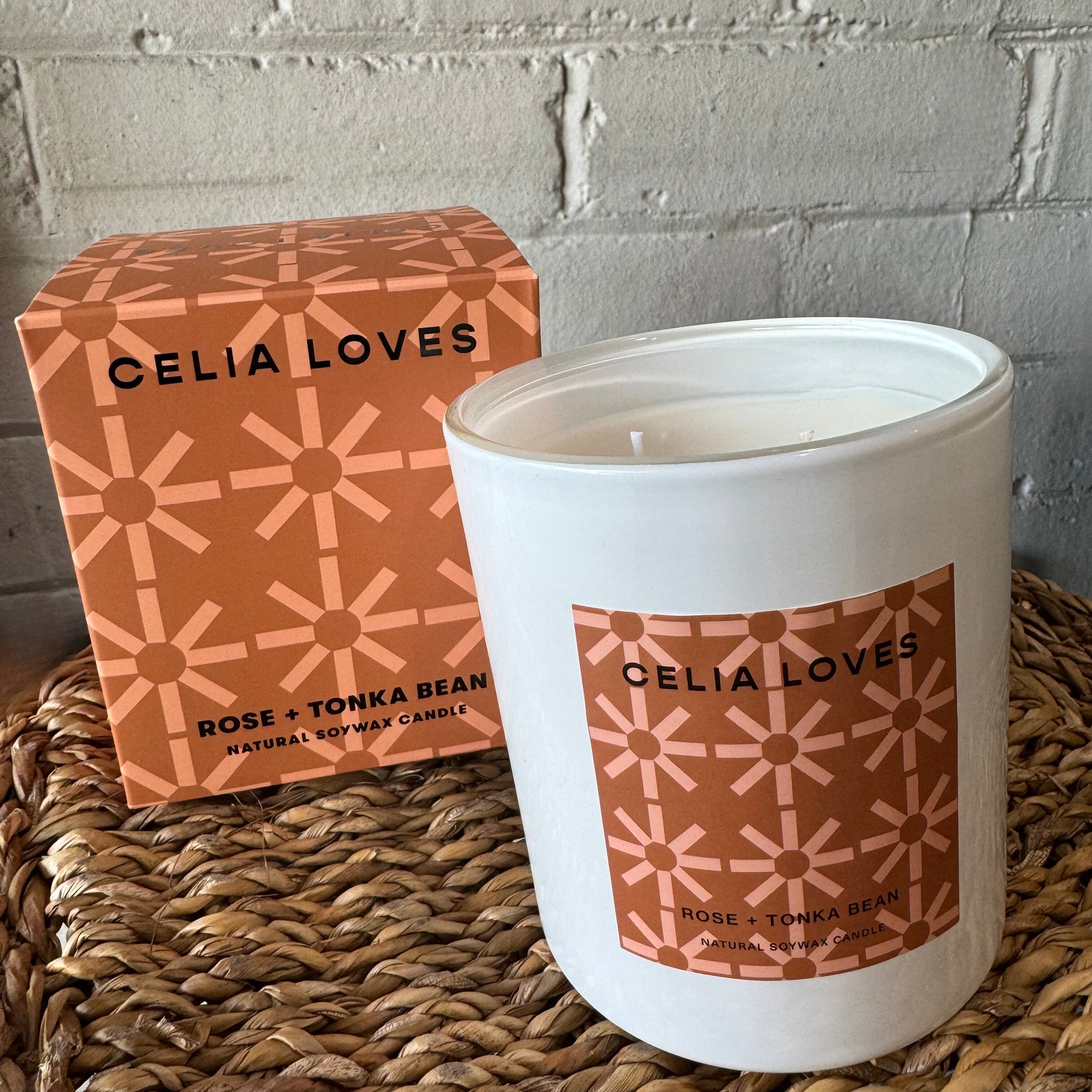 A candle from Flowers on Norton St's "Celia Loves Candles" collection, featuring the distinctive fragrance of "Rose + Tonka Bean," is displayed in a white glass jar next to its matching orange geometric-patterned box. These hand-poured candles are elegantly arranged on a woven surface against a backdrop of white brick.