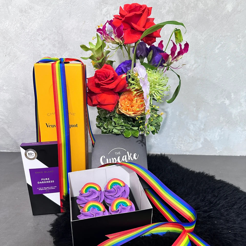 The Rainbow Pride arrangement from Flowers on Norton St showcases a vibrant display of multicolored flowers in a vase, complemented by a Veuve Clicquot champagne box, a chocolate box, and rainbow cupcakes adorned with purple frosting and rainbow toppers. A graceful rainbow ribbon drapes over the entire setup to celebrate Pride Month.