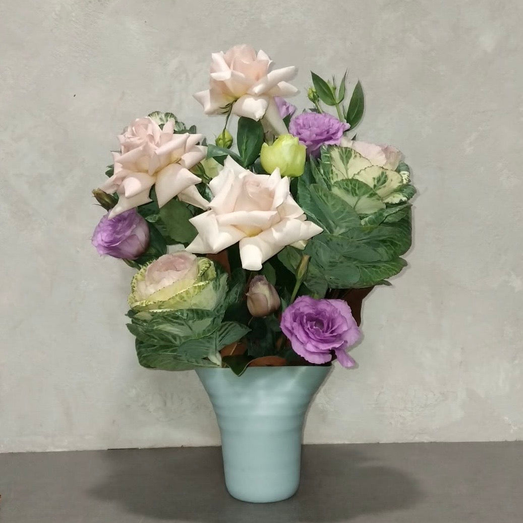 A floral arrangement featuring pale pink roses and purple lisianthus in a duck egg blue "Aurora Vase" by Flowers on Norton St, set against a plain background.