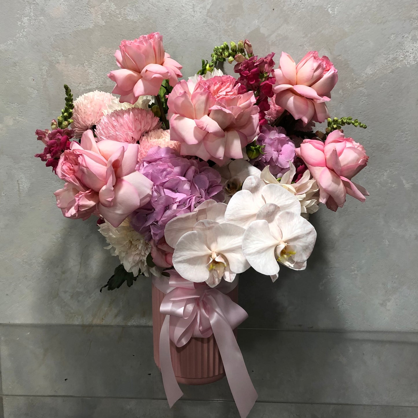 The Bubblegum Pop Pink Bouquet by Flowers on Norton St features pink roses, white orchids, purple hydrangeas, and seasonal flowers arranged in a pink ribbed vase with a ribbon against a light grey background.