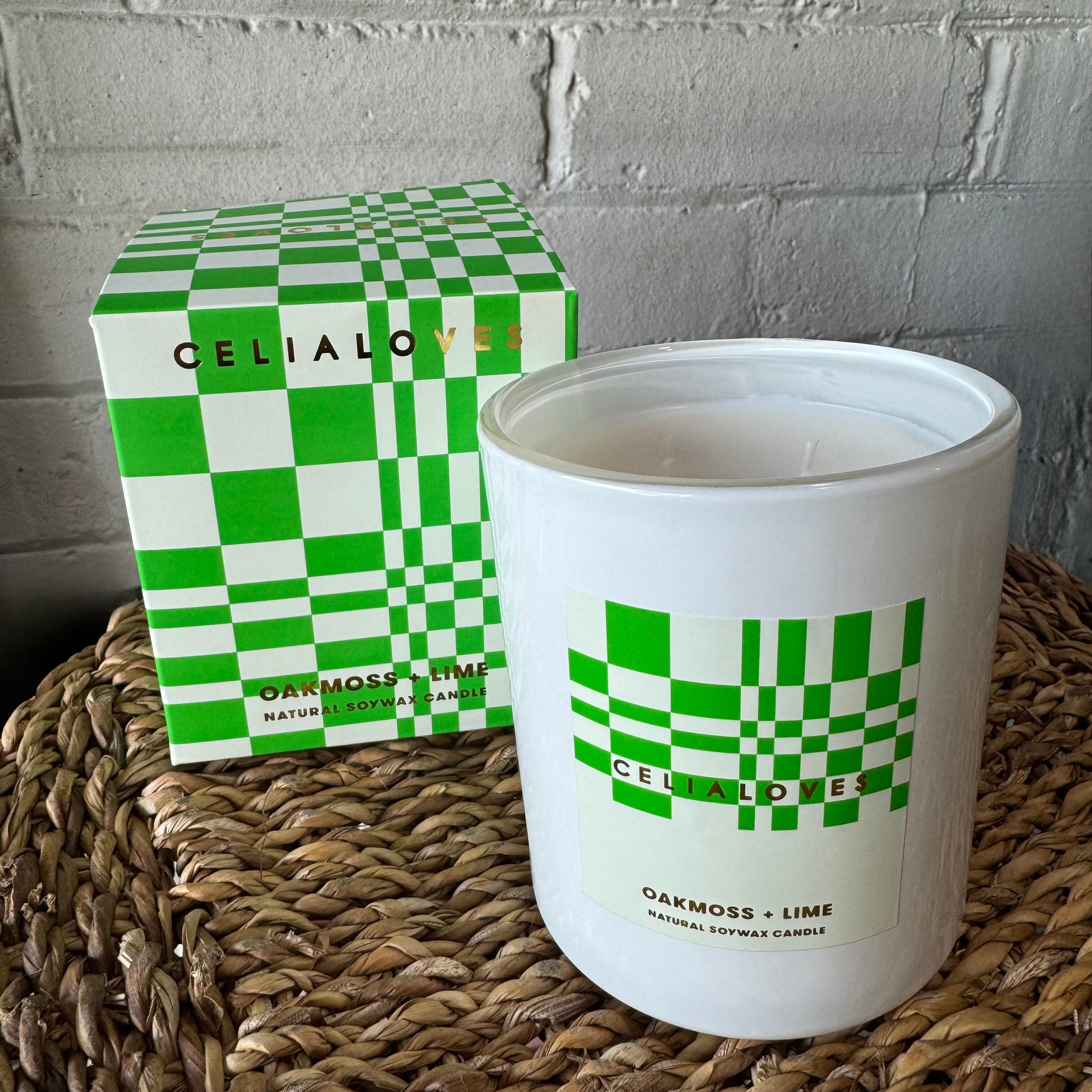 A hand-poured soy wax candle in white, labeled "OAKMOSS + LIME," with a green checkered design, sits alongside its matching box on a woven surface. The brand "Flowers on Norton St" offers distinctive fragrances for both the Celia Loves Candles and their packaging.