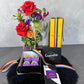 A vibrant display from the Rainbow Pride collection by Flowers on Norton St, featuring an array of colorful roses and mixed flowers in a vase. This is beautifully complemented by a box of rainbow Pride cupcakes adorned with rainbow toppers, a Veuve Clicquot champagne box, a chocolate bar, and accented with rainbow ribbons on a sleek black surface – capturing the joyful essence of Pride Month.