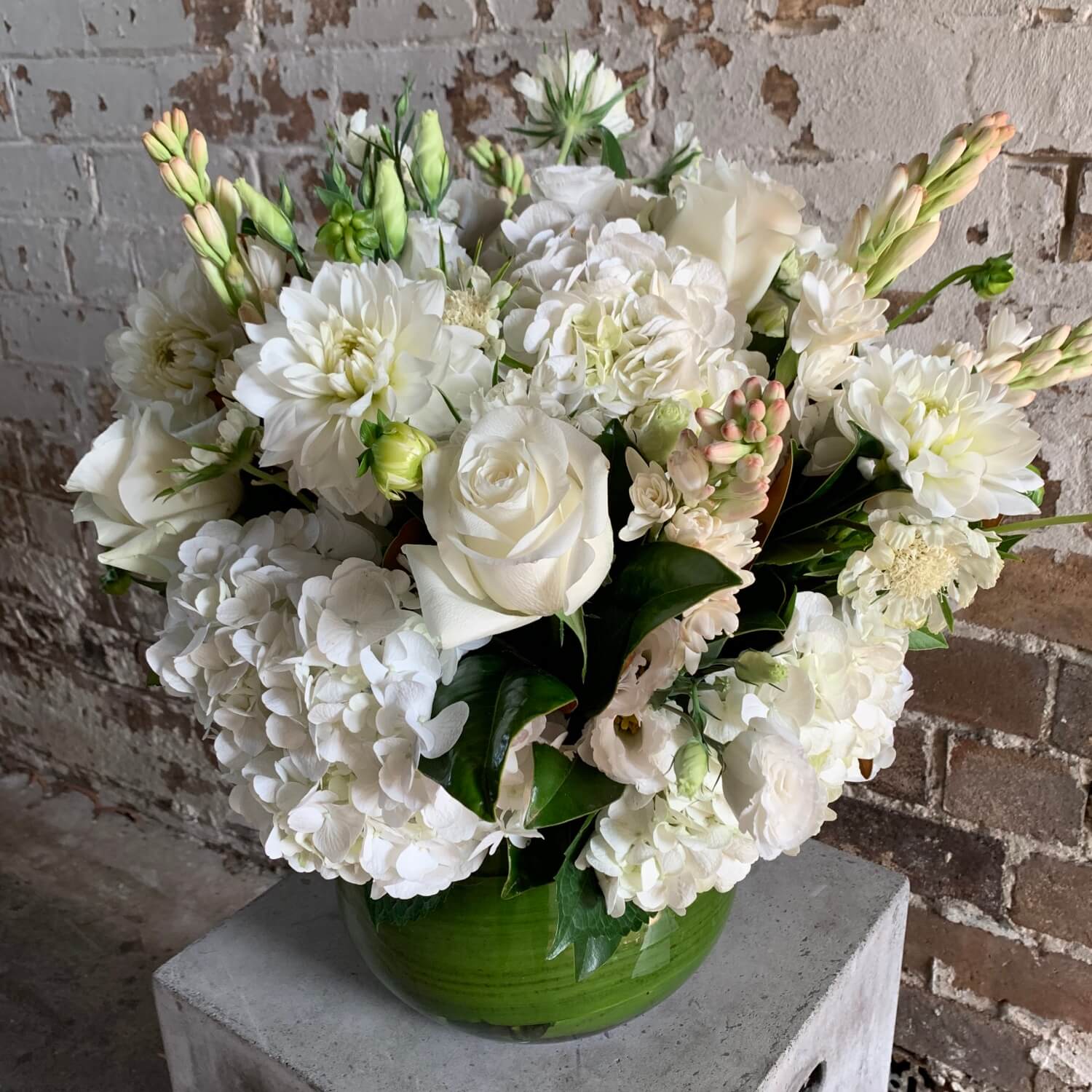 An elegant display of all white flowers, featuring roses and hydrangeas, is beautifully arranged in a green Divine Fleurs Vase by Flowers on Norton St. The vase is placed on a concrete block with a rustic brick wall as the backdrop.