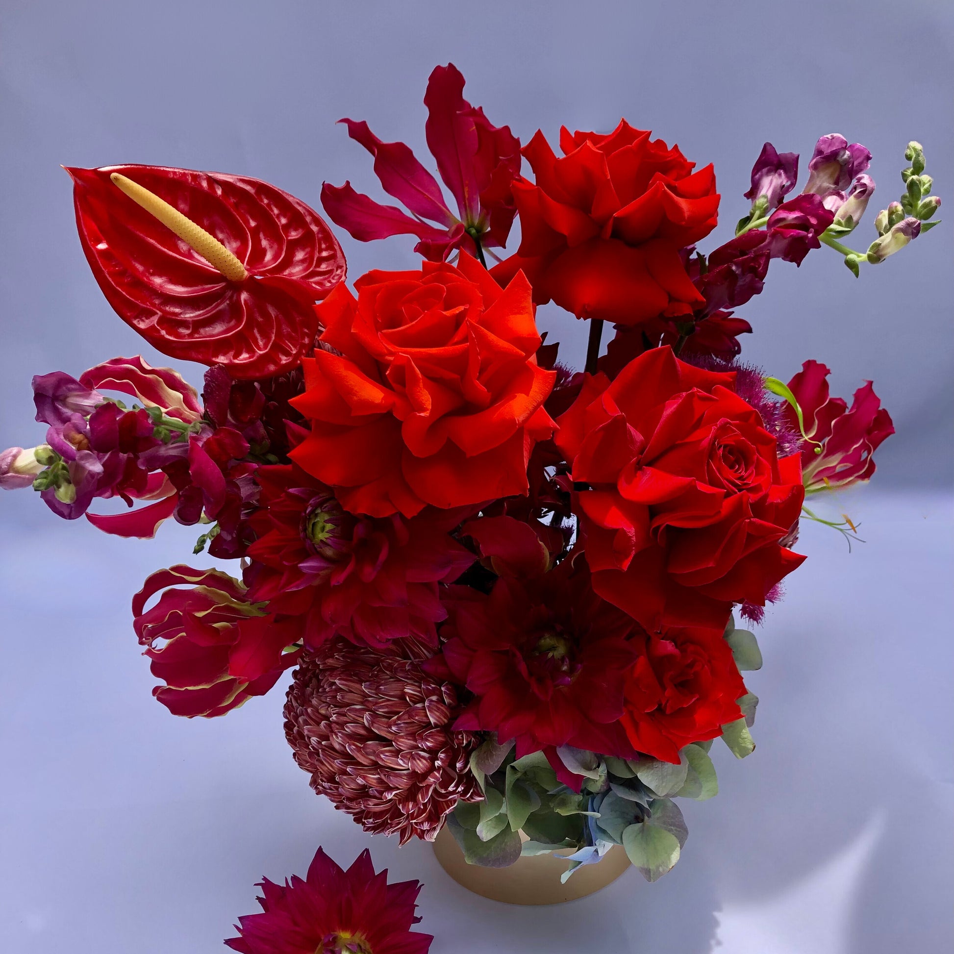 A vibrant floral arrangement showcasing red roses, anthuriums, and assorted red and burgundy blooms in a vase against a simple background. The petals and leaves gracefully cascade over the vase's edge, making it an ideal centerpiece for the A Soiree Affair Red Bouquet by Flowers on Norton St.