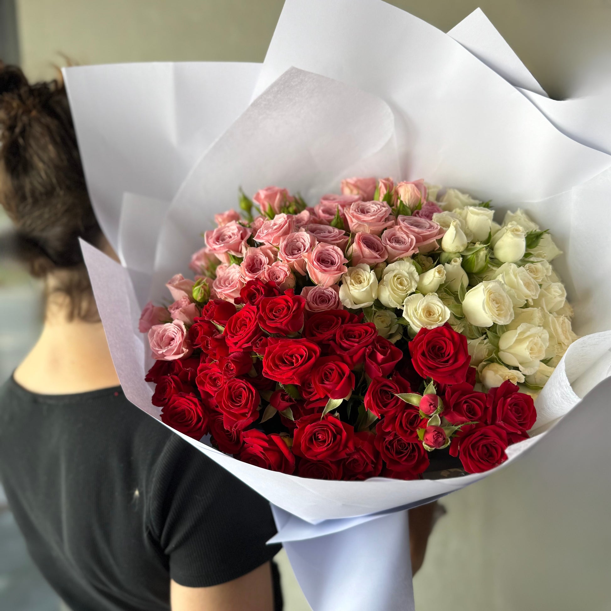 A person holds a stunning Mini Roses Enmasse Bouquet from Flowers on Norton St, elegantly wrapped in white paper against a gray backdrop. The bouquet's charm and romance shine as the individual, wearing a black shirt, faces away.