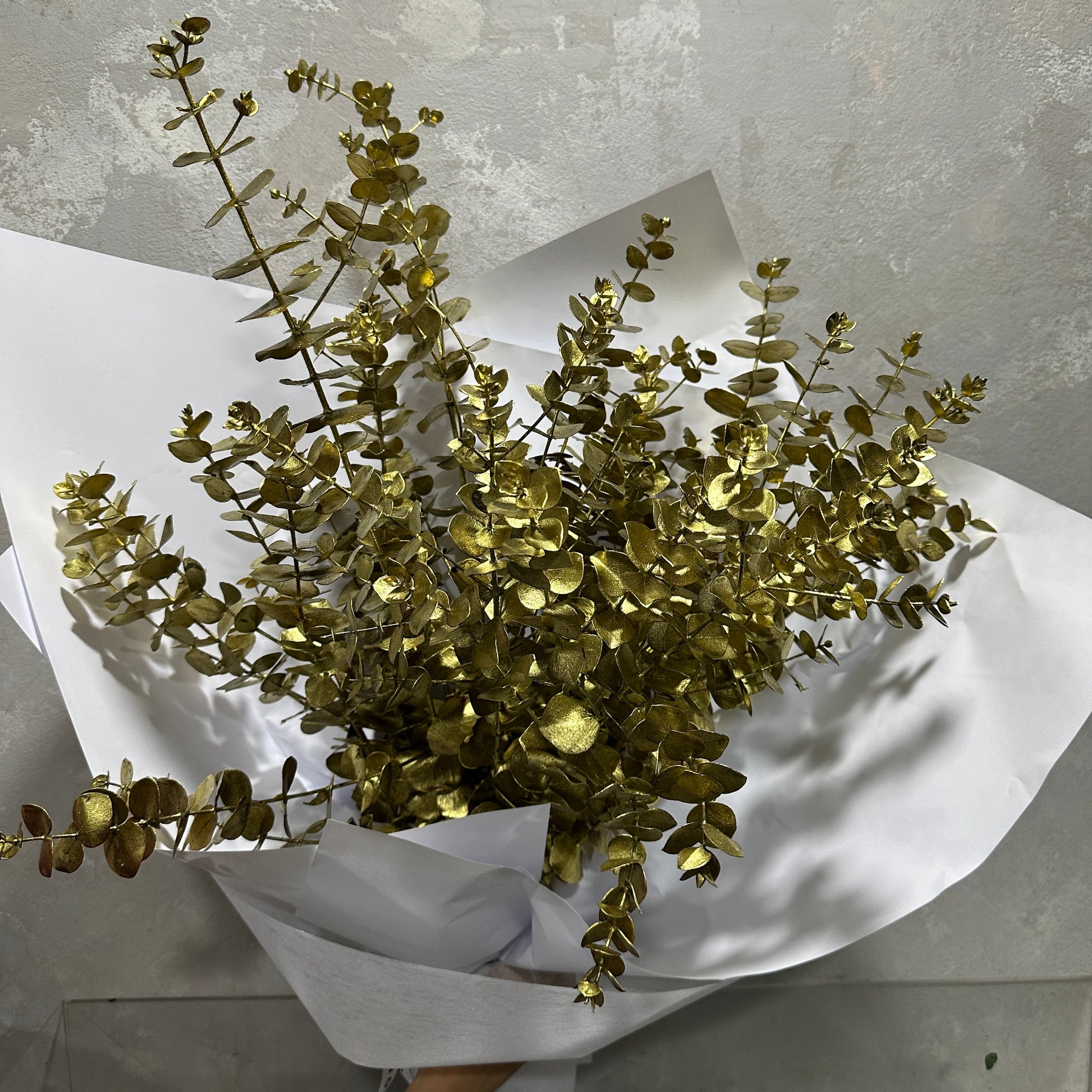 A bouquet of Flowers on Norton St's Dried Gold Xmas Foliage wrapped in white paper and set against a textured gray background.