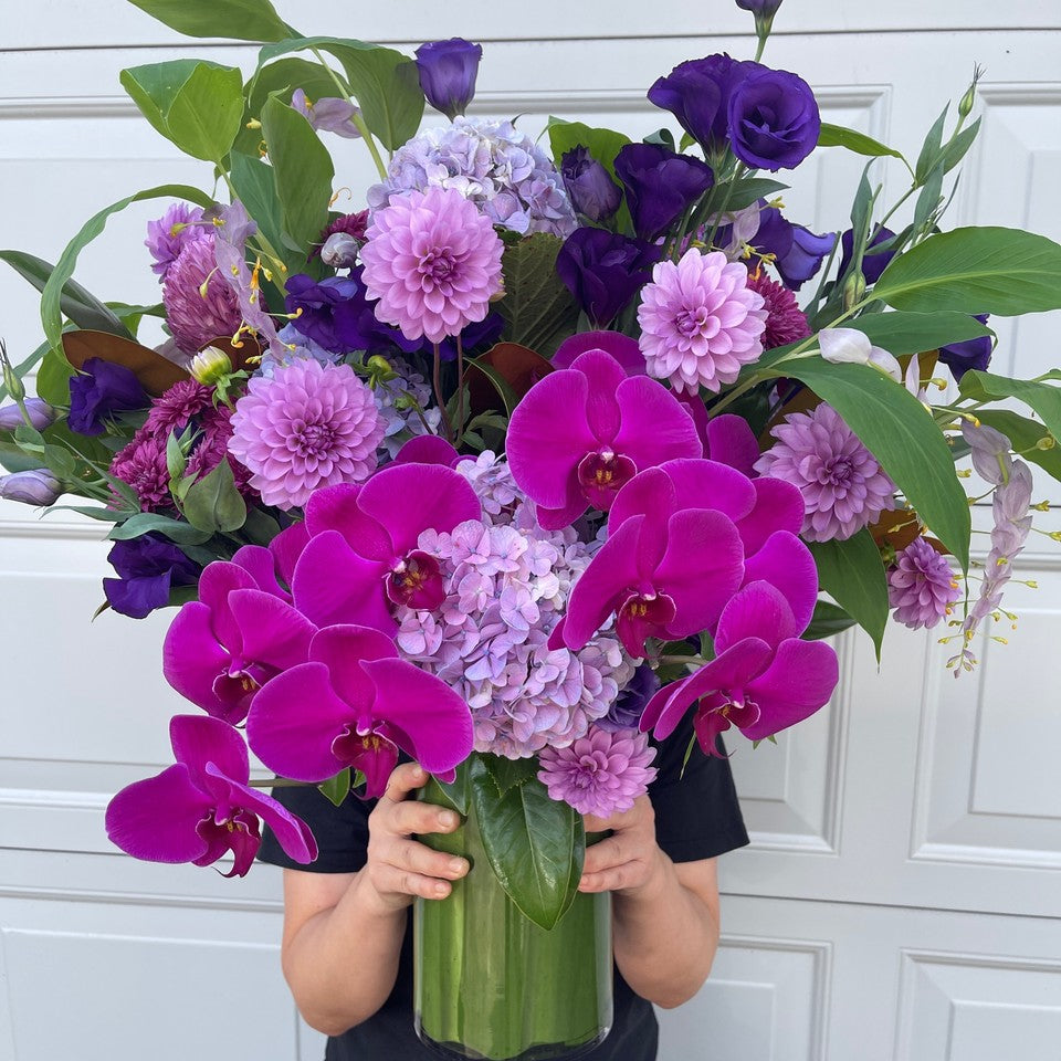 A person holds the "Parisian Purple" bouquet by Flowers on Norton St, showcasing vibrant blooms of phalaenopsis orchids and jewel-toned dahlias, beautifully complemented by lush green leaves. These flowers are elegantly arranged in a clear glass vase set against a white background, with the person's face hidden behind the eye-catching display.