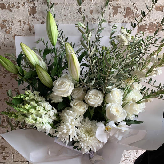 The FlowersonNortonSt Ivory Bliss Bouquet showcases an exquisite selection of white blooms, including roses, lilies, chrysanthemums, and snapdragons, beautifully complemented by rich greenery. Delicate orchids add an elegant touch against a backdrop of textured, weathered white brick wall.