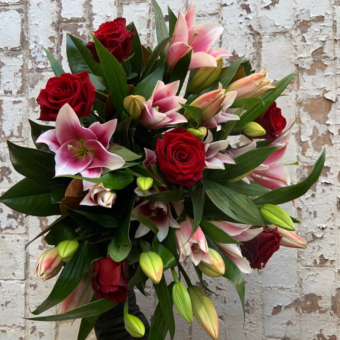 The Flowers on Norton St Red Rose + Pink Lily Bouquet, featuring red roses, pink lilies, and green leaves, sits elegantly against a textured white brick wall. With blooming lilies and buds, it's perfect for Valentine’s Day adding a fresh touch of color and contrast.