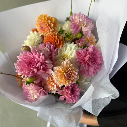 The Dahlia Delight - Summer Pastels Bouquet by Flowers on Norton St is perfect for weddings, featuring pink, orange, and white Dahlias wrapped in white tissue paper. The closely arranged ruffled petals create a vibrant display suitable for any celebration or special occasion.