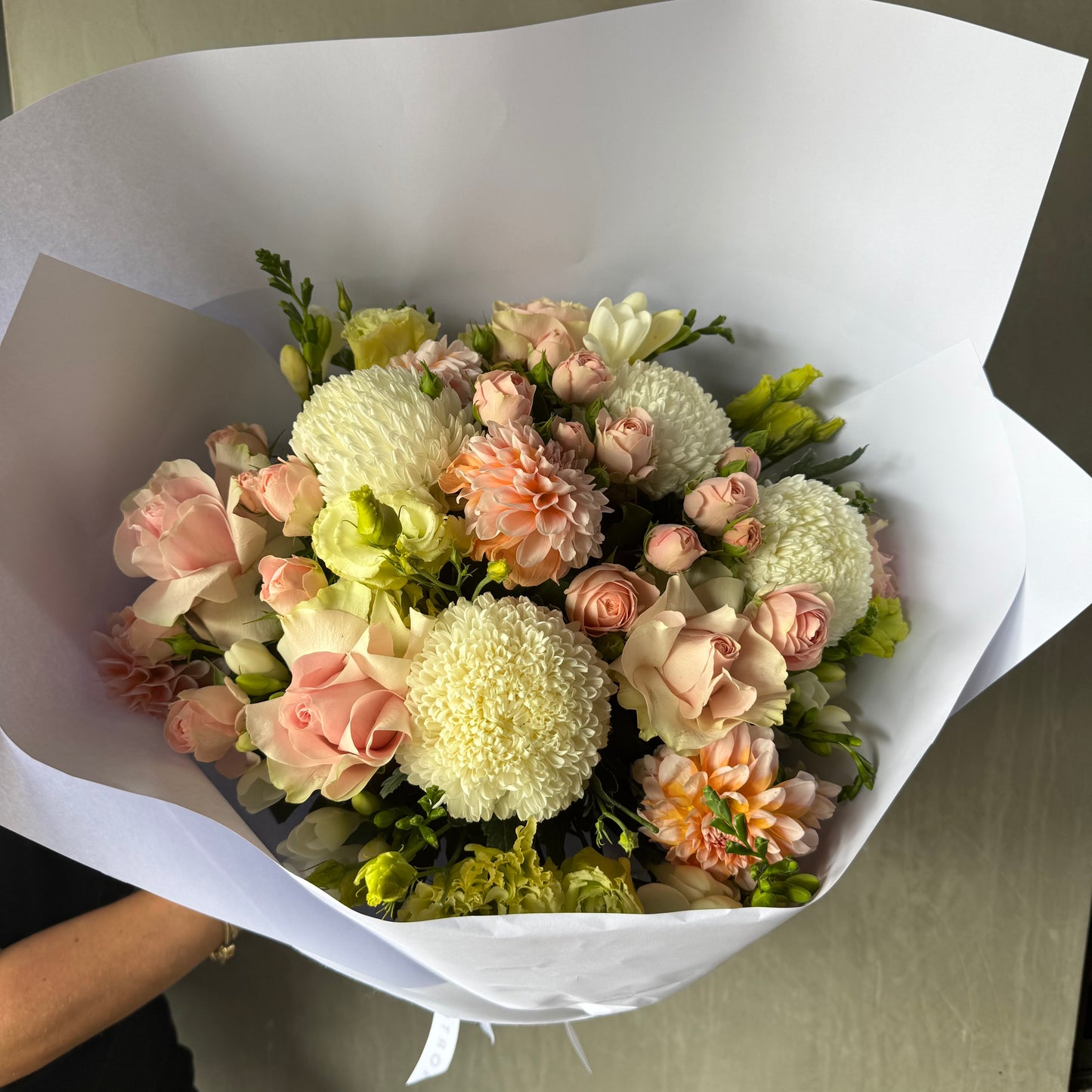 A Little Sweetheart Posy from Flowers on Norton St showcases in-season flowers, including pale pink roses, white and soft peach chrysanthemums, and greenery, wrapped in white paper and held against a neutral background.